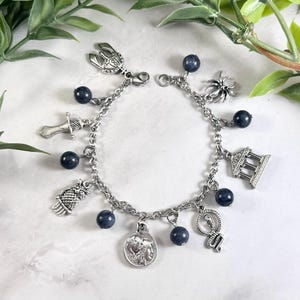 May include: A silver-toned charm bracelet featuring a series of dark blue spherical beads interspersed with various metal charms, including a helmet, a sword, an owl, a coin, a snake, a spider, and a classical building structure with columns.