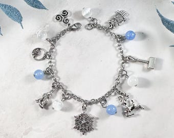 May include: A silver charm bracelet featuring various charms, including a snowflake, a reindeer, a hammer, and a wolf. The bracelet also has blue and white beads. The bracelet is displayed on a white surface with a blue decorative element.