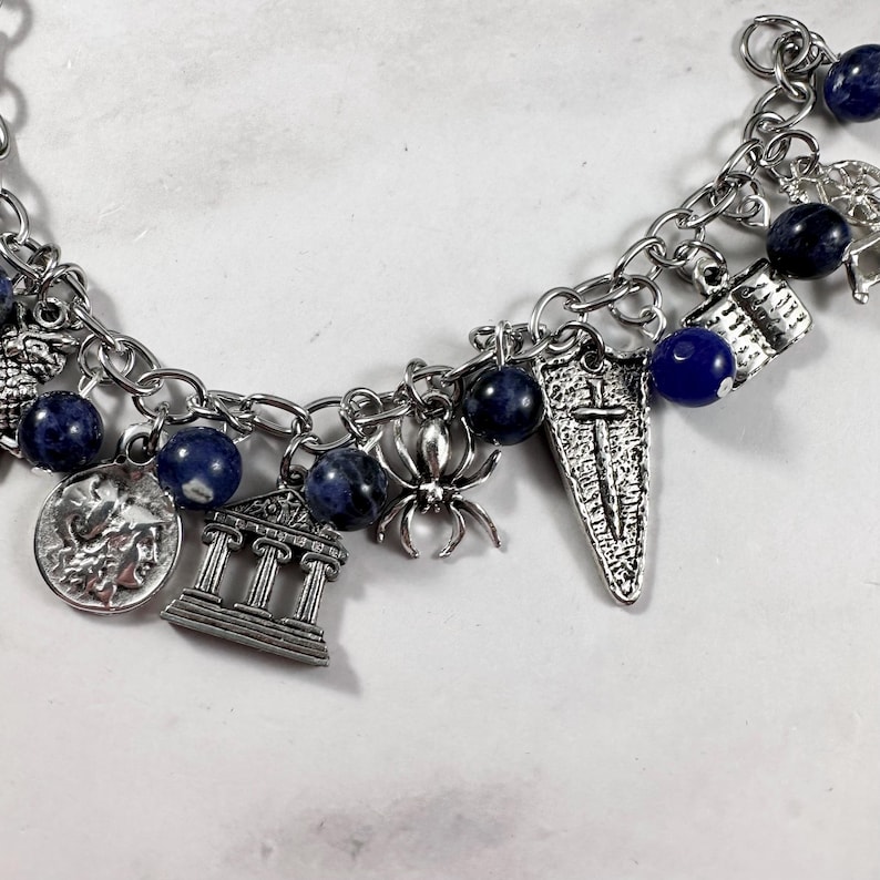 May include: A silver charm bracelet featuring blue beads and a variety of charms, including a spider, a temple, a shield, and a coin.