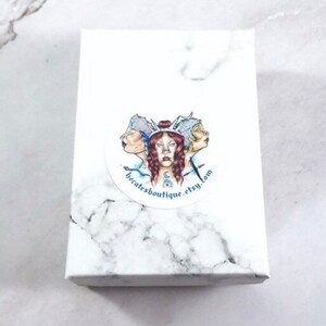 Puede incluir: Caja blanca con una pegatina redonda que presenta una ilustraci&oacute;n colorida de tres caras con una corona y el texto "becatesboutique.etsy.com".