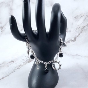 May include: A silver chain bracelet with black beads and charms, including a key, a circle, and a star. The bracelet is displayed on a black hand-shaped stand.