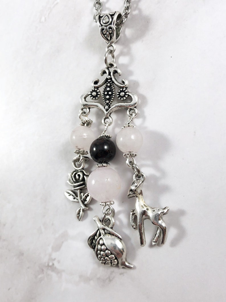 Puede incluir: Un collar de plata con un delicado colgante que presenta una rosa, un ciervo y un granado. El colgante est&aacute; adornado con cuentas rosas y negras.
