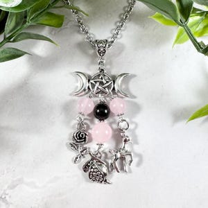 Puede incluir: Un collar de plata con un dije de triple luna y pentagrama, cuentas rosadas y negras, y dijes de plata de una rosa, un ciervo y una granada.