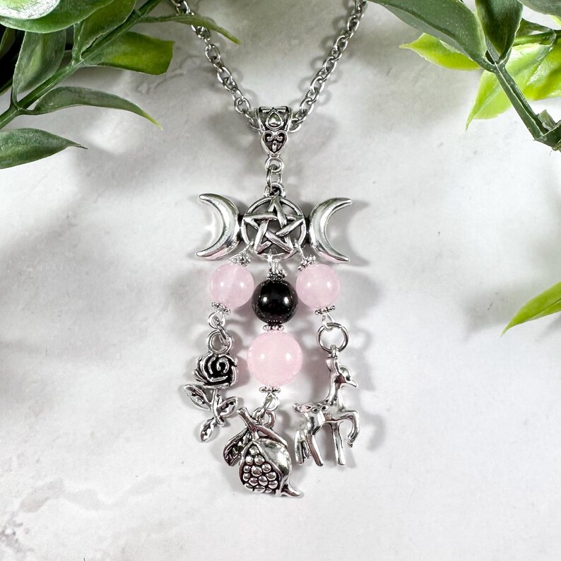Puede incluir: Un collar de plata con un dije de triple luna y pentagrama, cuentas rosadas y negras, y dijes de plata de una rosa, un ciervo y una granada.