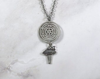 Puede incluir: Un collar de plata con un colgante circular que presenta un diseño de laberinto y un colgante más pequeño con forma de árbol estilizado con un pájaro en la parte superior.