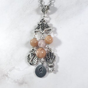 May include: Silver charm necklace with a sunstone, rose quartz, and a silver charm of a swan, wheat, and a rune.