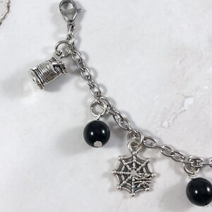 May include: A silver charm bracelet with black beads, a spiderweb charm, and a spool of thread charm.