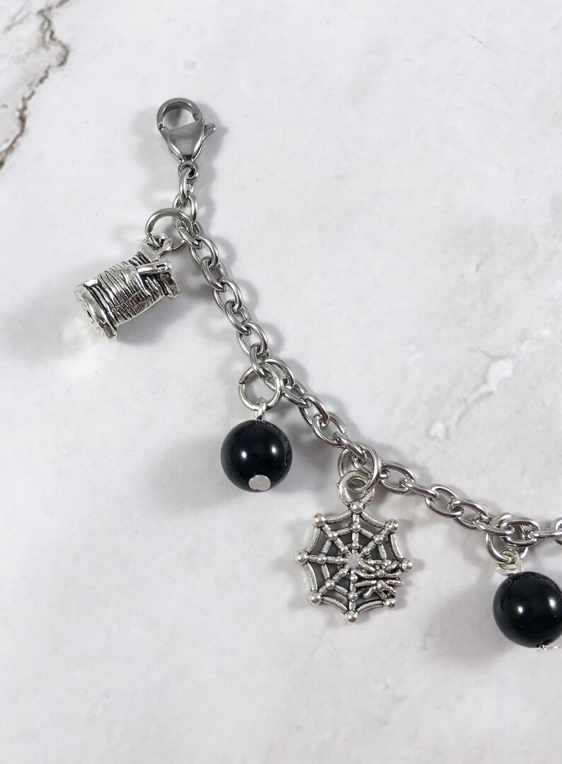 May include: A silver charm bracelet with black beads, a spiderweb charm, and a spool of thread charm.