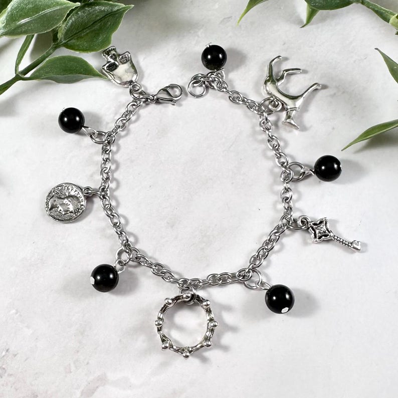 May include: A silver chain bracelet with multiple charms, including a skull, a deer, a coin, a key, and a crown. The bracelet also features black beads.