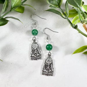Puede incluir: Pendientes plateados con una cuenta verde y un colgante que representa un faraón egipcio. Los pendientes tienen cierre de gancho y se muestran sobre un fondo blanco con follaje verde.