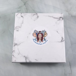 May include: A white marble box with a round sticker featuring a colorful illustration of three women with blue and white clothing. The sticker also includes the text "becatesboutique.etsy.com".