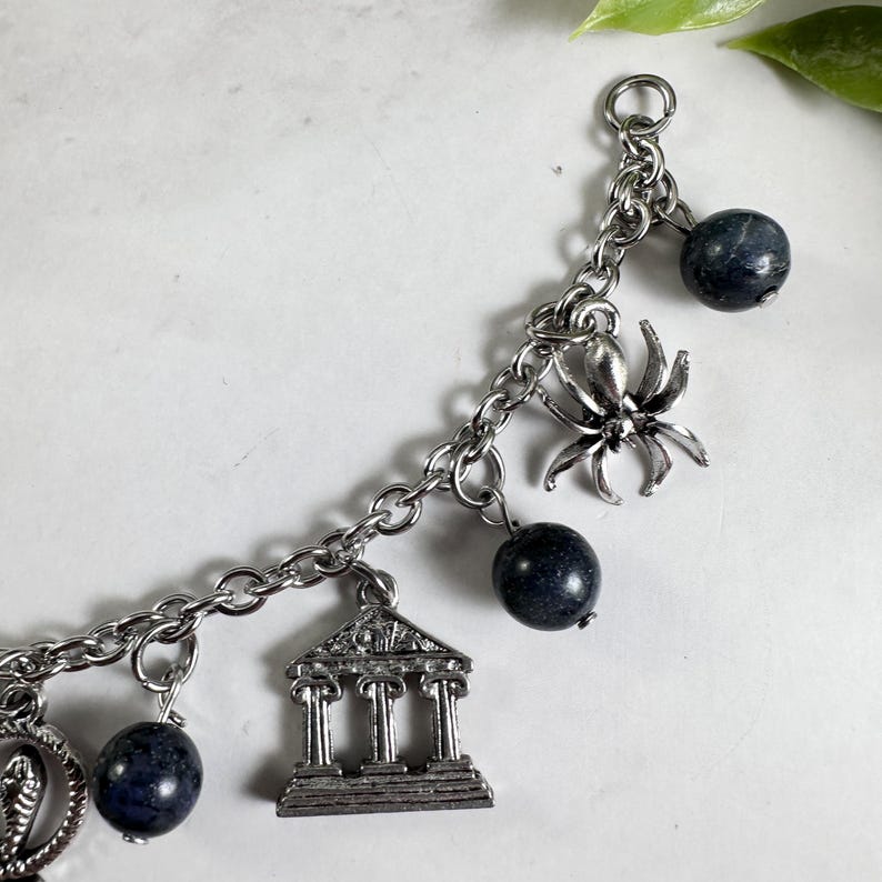 May include: A silver-toned metal charm bracelet featuring a series of dangling charms including a detailed spider, a classical building with columns, and a circular snake motif, interspersed with round, dark blue, mottled stone beads.