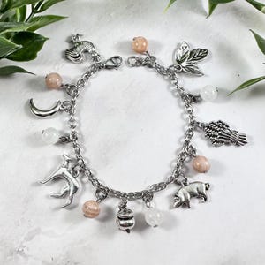 May include: A silver charm bracelet with a variety of charms, such as a bird, a crescent moon, a dog, a pig, and a pot. The bracelet also features pink and white gemstone beads. The bracelet is displayed on a white surface with green foliage.