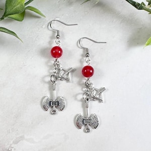 May include: A pair of drop earrings featuring silver-toned metal charms shaped like a double-headed battle axe and a bow and arrow. Each earring includes a single round, smooth red bead suspended from a silver-toned metal fishhook ear wire.