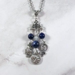 May include: A silver necklace with a pendant featuring a silver owl, snake, and coin with a face on it. The pendant is attached to a silver chain with blue and white beads.