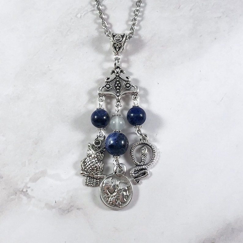 May include: A silver necklace with a pendant featuring a silver owl, snake, and coin with a face on it. The pendant is attached to a silver chain with blue and white beads.