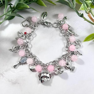 Puede incluir: Una pulsera de plata con abalorios de piedras preciosas rosas y varios dijes. Los dijes incluyen un corazón, arco y flecha, rosa, espejo, paloma y una cara. La pulsera se muestra sobre una superficie blanca con follaje verde.