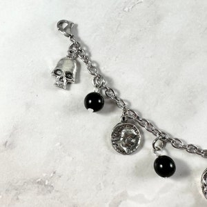 May include: A silver charm bracelet with black beads, a skull charm, and a coin charm. The bracelet is on a white background.