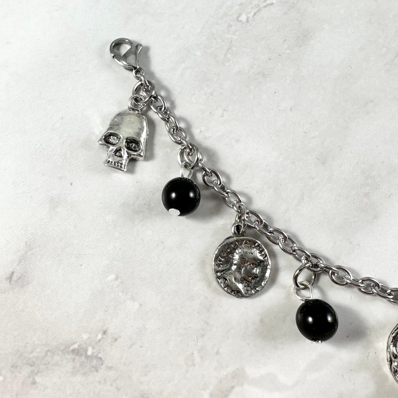 May include: A silver charm bracelet with black beads, a skull charm, and a coin charm. The bracelet is on a white background.