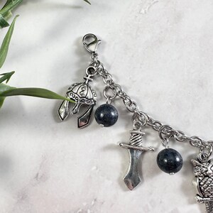 May include: A silver-toned charm bracelet featuring a series of dangling charms including a detailed helmet, a sword, and an owl, interspersed with round, dark grey speckled beads. The charms are attached to a silver-toned metal link chain.