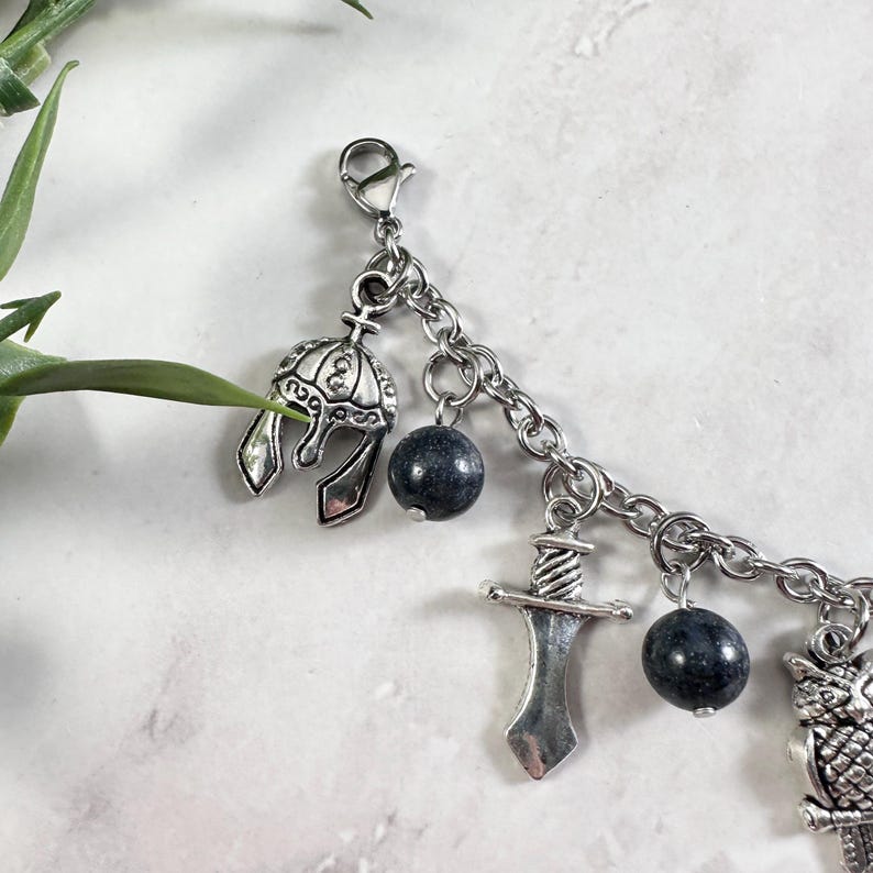 May include: A silver-toned charm bracelet featuring a series of dangling charms including a detailed helmet, a sword, and an owl, interspersed with round, dark grey speckled beads. The charms are attached to a silver-toned metal link chain.
