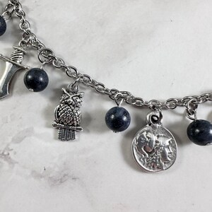 May include: A silver-toned charm bracelet featuring a series of dark blue, spherical beads interspersed with metal charms. The charms include a detailed owl, a small sword, and a circular coin pendant depicting a profile of a classical figure.