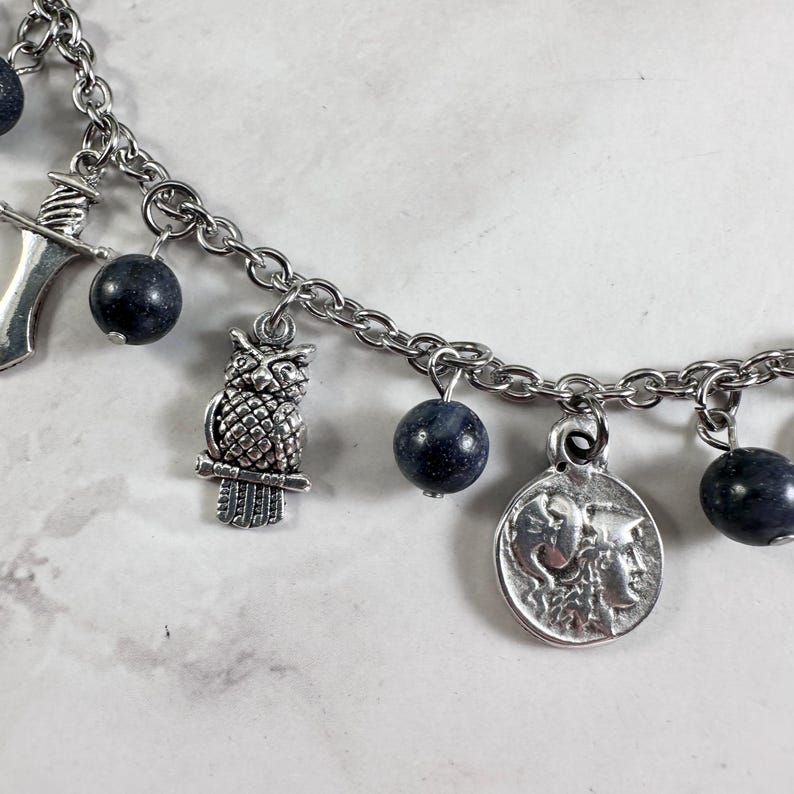May include: A silver-toned charm bracelet featuring a series of dark blue, spherical beads interspersed with metal charms. The charms include a detailed owl, a small sword, and a circular coin pendant depicting a profile of a classical figure.