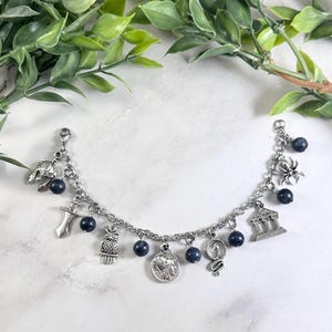 May include: A silver-toned charm bracelet featuring a series of dangling charms including a helmet, sword, owl, coin, snake, building, and spider, interspersed with small, round, dark blue beads attached to a classic metal link chain.