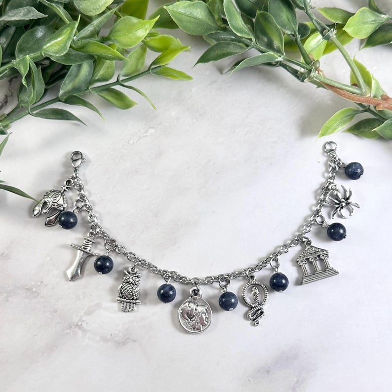 May include: A silver-toned charm bracelet featuring a series of dangling charms including a helmet, sword, owl, coin, snake, building, and spider, interspersed with small, round, dark blue beads attached to a classic metal link chain.
