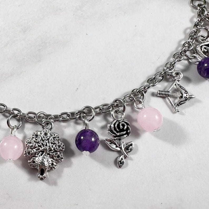 May include: A silver charm bracelet with pink and purple beads, a silver tree charm, a silver rose charm, and a silver bow and arrow charm.