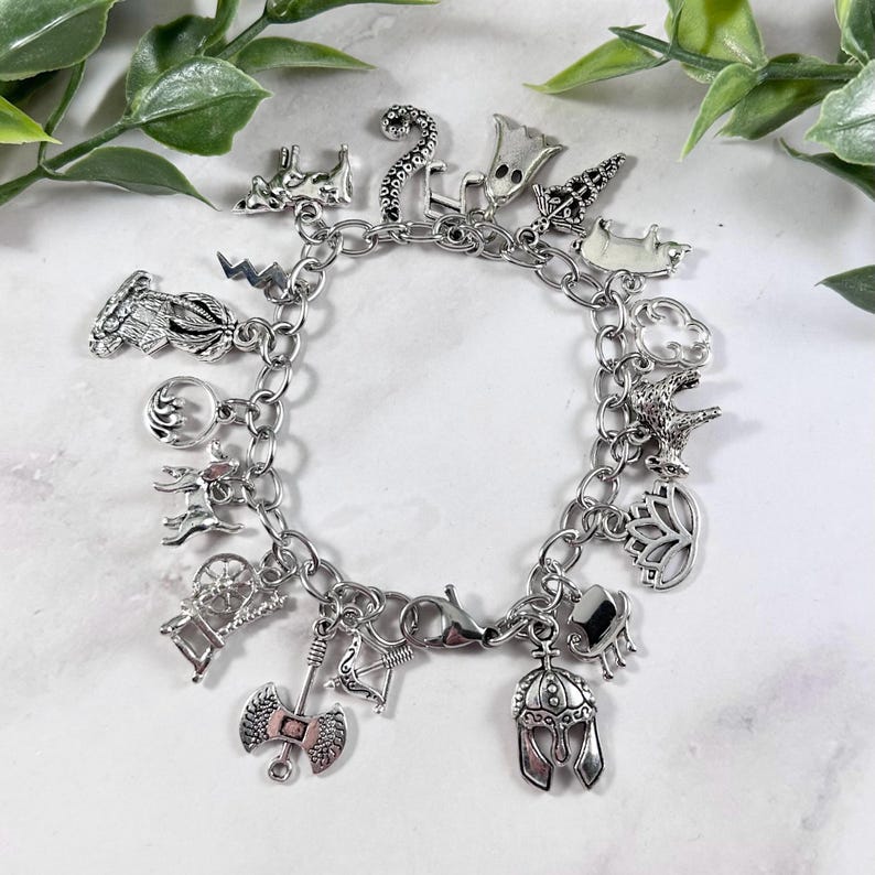 May include: A silver charm bracelet featuring a variety of charms, including a crown, a crescent moon, a lotus flower, a spinning wheel, an axe, a bow and arrow, a helmet, a horse, a pig, a ghost, a lightning bolt, a snake, a deer, and a clover.
