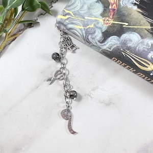 May include: A silver bookmark chain with charms, including a spinning wheel, scissors, a measuring tape, and black beads. The bookmark is hanging from a book titled "Greek Myths".