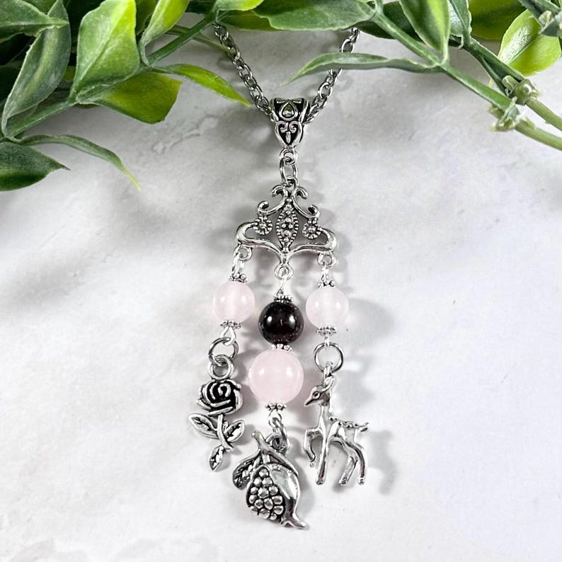 Puede incluir: Un collar de plata con un delicado colgante que representa una rosa, un ciervo y una granada. El colgante est&aacute; adornado con cuentas rosas y negras.
