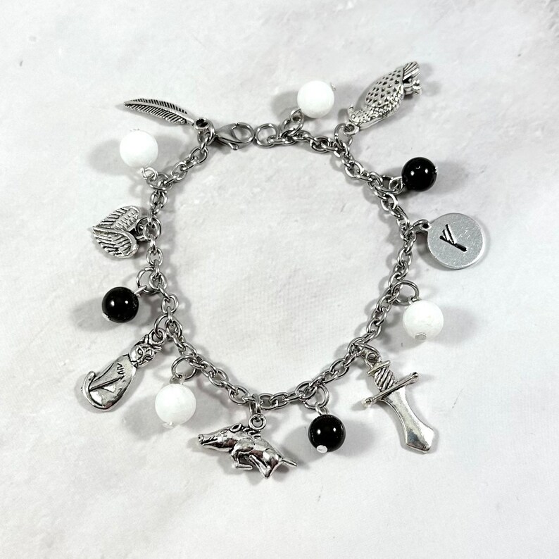 May include: A silver charm bracelet with various charms, including a feather, a heart with wings, a boar, a sword, a bird, a rune, and white and black beads.