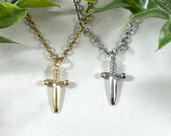 Puede incluir: Dos collares con colgantes de daga. Uno tiene una cadena y un dije dorados, el otro una cadena y un dije plateados. Las dagas tienen mangos y hojas detallados.