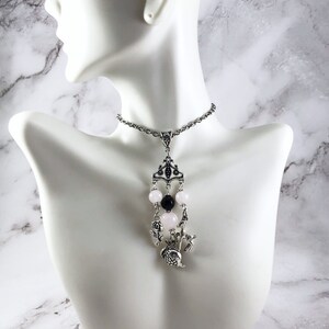 Puede incluir: Un collar de cadena de plata con un colgante con un dise&ntilde;o de corona, cuentas rosas y negras y un peque&ntilde;o amuleto de p&aacute;jaro.