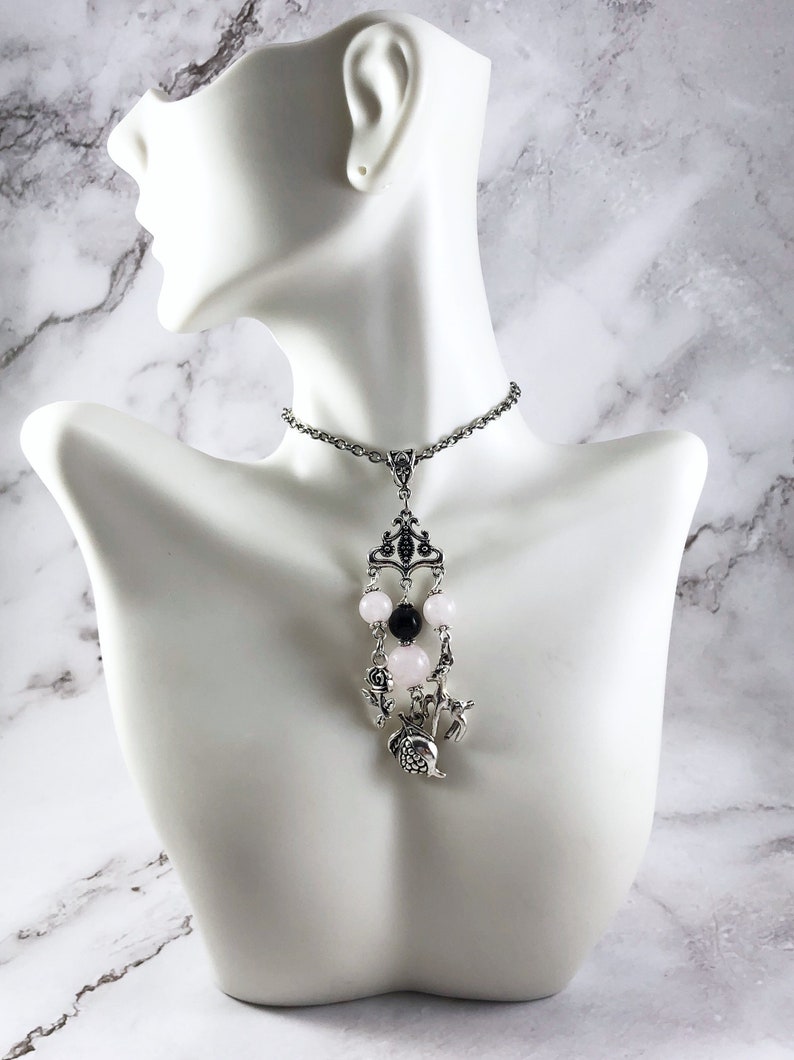 Puede incluir: Un collar de cadena de plata con un colgante con un dise&ntilde;o de corona, cuentas rosas y negras y un peque&ntilde;o amuleto de p&aacute;jaro.