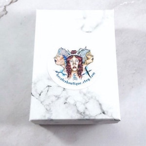 May include: White box with a round sticker featuring a drawing of three women with blue and white crowns. The sticker has the text "becatesboutique.etsy.com".
