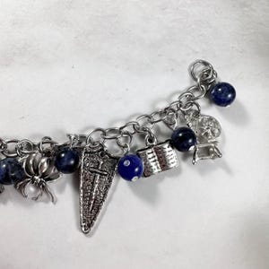 May include: A silver charm bracelet featuring blue beads and a variety of charms, including a spider, a book, a shield, a spinning wheel, and a dice.