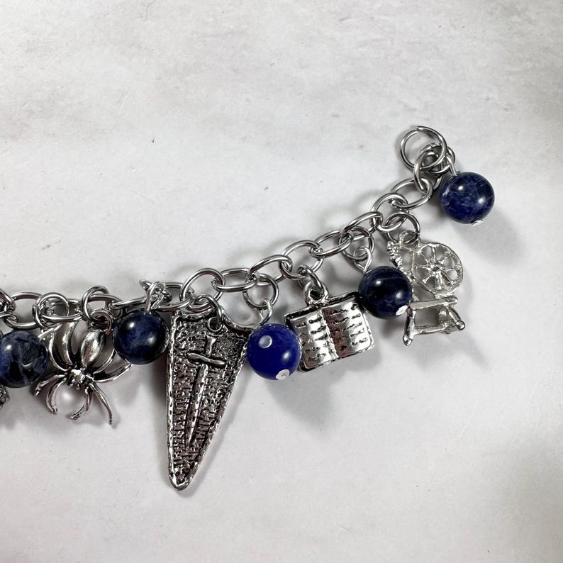 May include: A silver charm bracelet featuring blue beads and a variety of charms, including a spider, a book, a shield, a spinning wheel, and a dice.