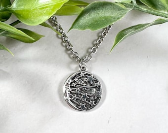 Puede incluir: Un collar de plata con un colgante redondo que presenta una luna creciente y estrellas. El colgante está detallado con una superficie texturizada.