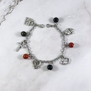 May include: A silver charm bracelet with a variety of charms, including a skull, a cross, a cloud, a building, and an eagle. The bracelet also features black and red beads.