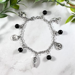 Puede incluir: Un brazalete de plata con cuentas negras y varios dijes de plata, incluyendo una calavera, un barco, una ola y una cara.