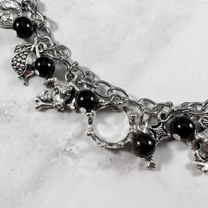 May include: Silver-toned charm bracelet featuring various charms, including a goat, a pomegranate, and a round charm. The bracelet also has black, round beads. The chain is made of silver-colored metal.