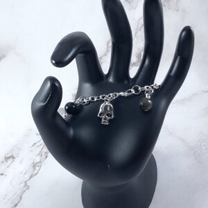 May include: A silver chain bracelet with a black bead, a silver skull charm, and a green stone charm.
