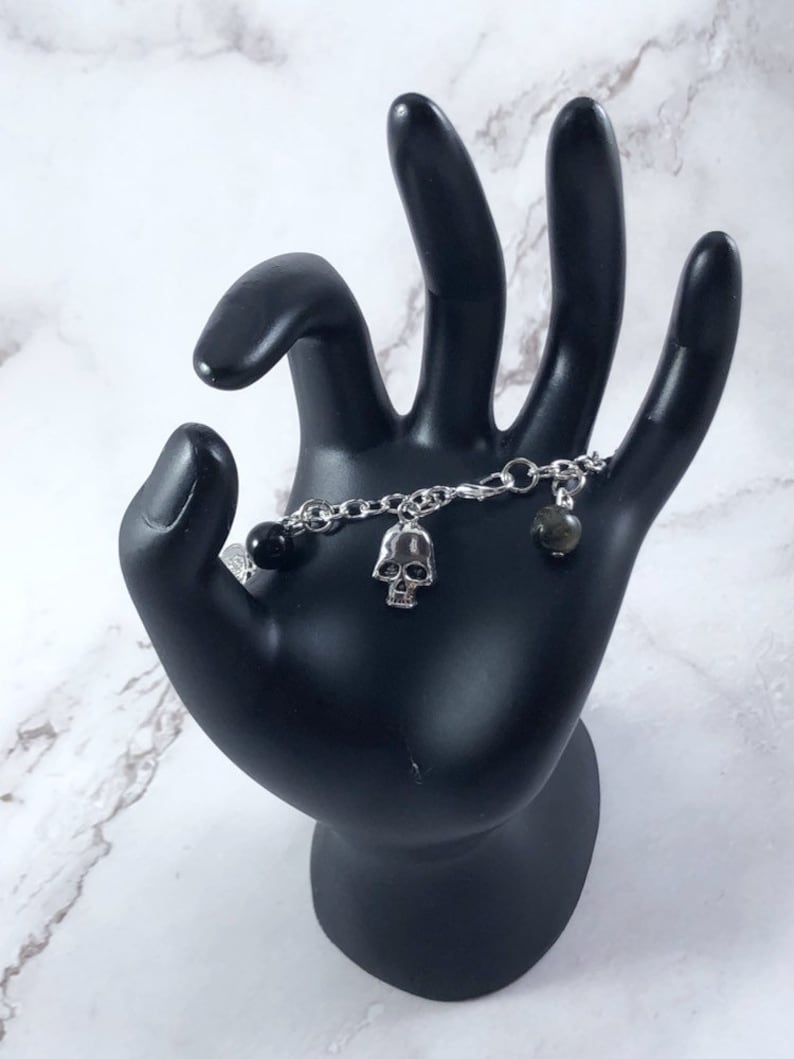 May include: A silver chain bracelet with a black bead, a silver skull charm, and a green stone charm.