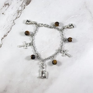 May include: A silver charm bracelet with brown tiger's eye beads, an Egyptian cat charm, an ankh charm, and a hand charm.