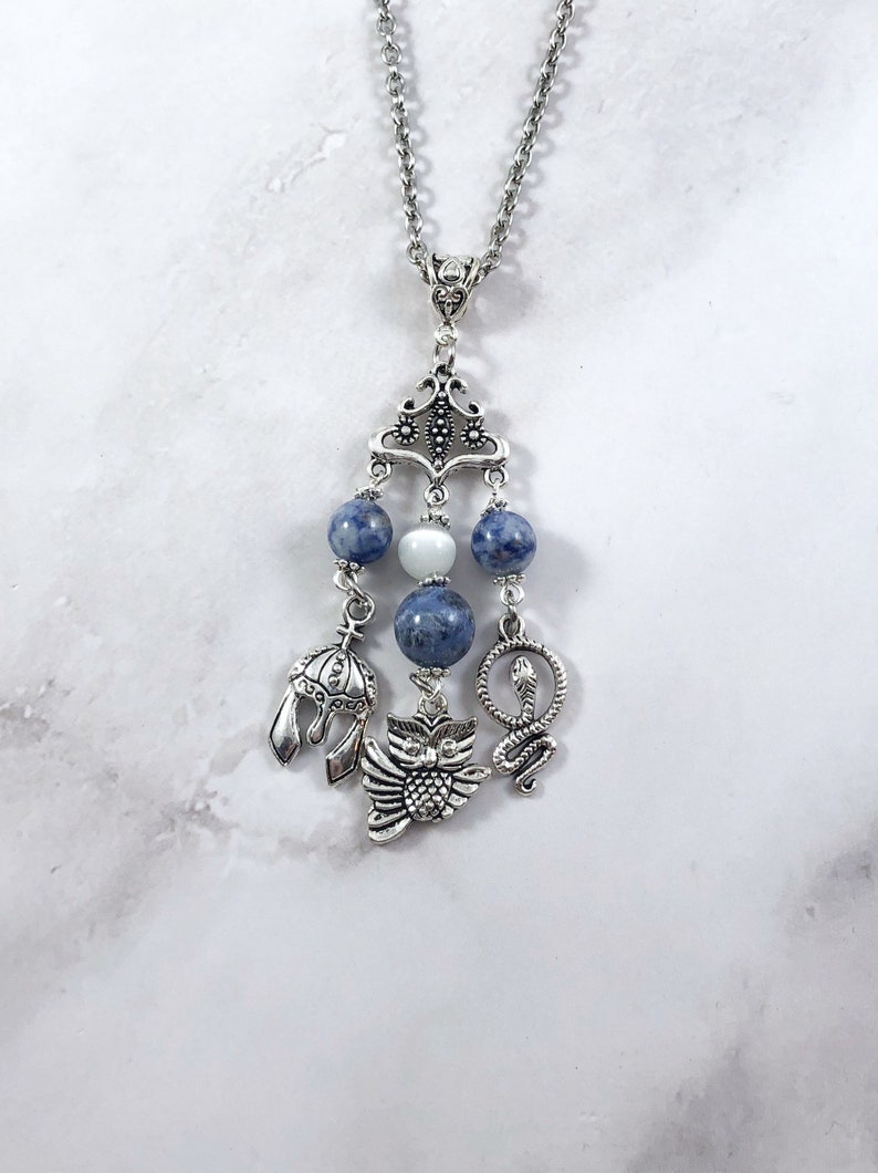May include: A silver necklace with a pendant featuring a silver owl, snake, and helmet charm. The pendant is adorned with blue and white beads.