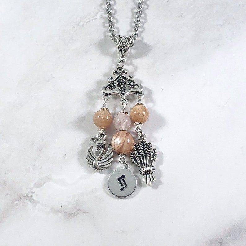May include: A silver chain necklace with a pendant featuring a silver charm with a swan design, a charm with wheat stalks, and a charm with the rune Algiz. The pendant is adorned with four peach-colored beads.