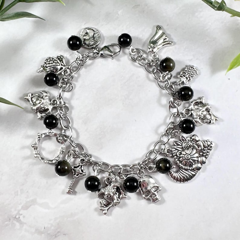 May include: Silver charm bracelet featuring various charms, including skulls, a key, and a coin. The bracelet also has black beads interspersed throughout the chain. The charms are detailed and the bracelet is on a white surface.